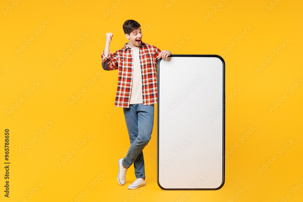 Poster Full body young man he wears red checkered shirt casual clothes big huge blank screen mobile cell phone smartphone with area do winner gesture isolated on plain yellow background. Lifestyle concept.