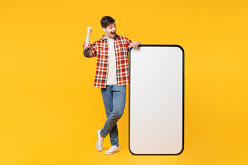 Full body young man he wears red checkered shirt casual clothes big huge blank screen mobile cell phone smartphone with area do winner gesture isolated on plain yellow background. Lifestyle concept.