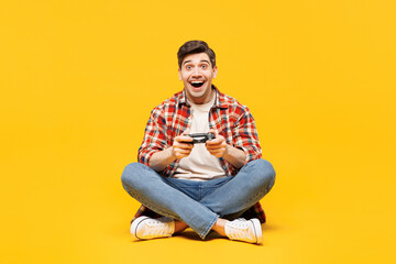 Full body young excited man wears red checkered shirt casual clothes sits hold in hand play pc game with joystick console isolated on plain yellow orange background studio portrait. Lifestyle concept.