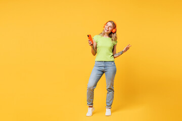 Fototapeta premium Full body young woman she wear green t-shirt casual clothes listen to music in headphones hold in hand use mobile cell phone look aside isolated on plain yellow orange background. Lifestyle concept.