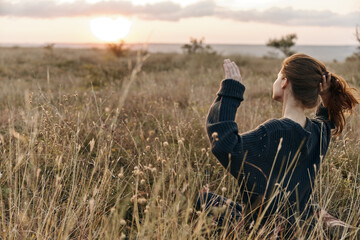 Woman sitting in golden field, hands raised to the sky at sunset, feeling connected to nature