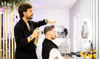 An adult hairdresser with a beard and black clothes is using a can of spray hairspray on a client's...