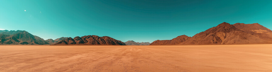 Wide-angle view of a barren desert landscape with cracked soil, leading to rocky mountains under a clear blue sky, suggesting arid conditions and open space.