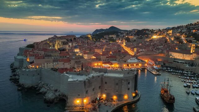 Dubrovnik old town after sunset with city lights. Aerial shot of historic city of Dubrovnik in Croatia, UNESCO World Heritage site. Illuminated streets, buildings and marina with boats. 4K HDR shot 