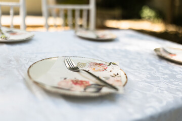Elegant dinner setting with floral plates and silverware arranged on a white tablecloth in the afternoon sunlight