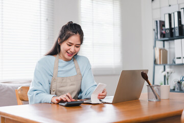 A woman in light blue sweater and apron smiles while using calculator and reviewing receipt at wooden table with laptop. Her cheerful expression adds warmth to workspace.