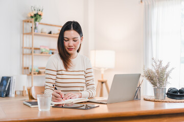 A woman is sitting at wooden desk, writing in notebook with laptop nearby. bright, airy room features shelf with decorative items and plant, creating calm workspace.