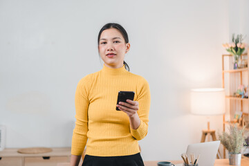 A young woman in yellow turtleneck sweater stands confidently holding smartphone in modern workspace, exuding sense of professionalism and focus.