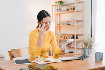 A woman in yellow sweater is sitting at wooden desk, looking stressed while holding notepad and pen. She is surrounded by laptop, books, and stationery, creating focused work environment.