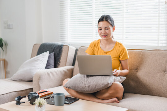 A young woman sits comfortably on sofa, working on her laptop with cheerful expression. cozy living room features natural light and relaxed atmosphere, perfect for productivity.