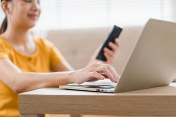Fototapeta premium A woman in yellow shirt is using laptop while holding smartphone, showcasing modern work from home setup. Her focused expression reflects productivity and multitasking.
