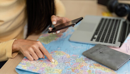 Young Woman Checking Travel Information on Smartphone with Map and Passport on Desk, Planning Vacation Trip