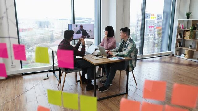 Team collaborating with remote colleague through video conference. Office setting with post-it notes on glass, laptops, and discussion. Modern workspace.