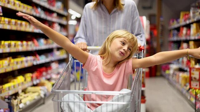 Little girl having fun riding in a shopping cart while shopping groceries at the supermarket with her mother