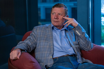 Senior man dressed in a blazer and jeans sits pensively in a stylish red chair, surrounded by...