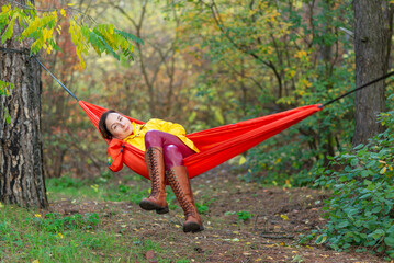 A young woman in a yellow raincoat and leather pants poses in an autumn forest while lying in a hammock