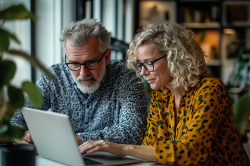 A middle-aged man and woman in an office, discussing business ideas while sitting at the table with a laptop computer near them.