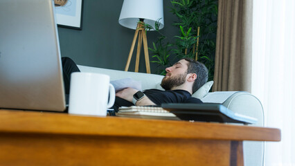 A man in a black shirt is lying on a couch, taking a nap during a work break. A laptop, mug, and papers are on the table in the foreground, moment of relaxation amidst a busy day.