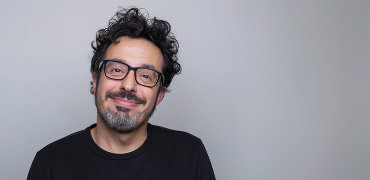 Studio headshot portrait of smiling friendly Jewish man with facial hair, glasses, and black cotton t shirt, gray background