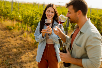 A joyful couple enjoying wine tasting together in a sunny vineyard during the golden hour,...