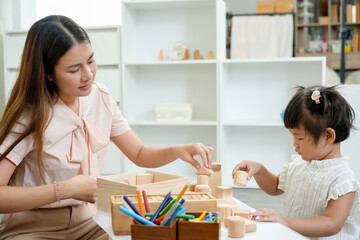 Fototapeta premium Asian woman sitting at a table playing with wooden blocks alongside toddler. early education, building motor skills and cognitive development. Calm and focused atmosphere in cozy home setting.