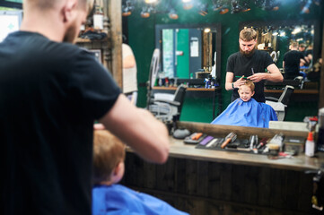 Little boy annoyed by hair falling on his eyes. Blond kid looking in the mirror and sad about new haircut. Little client sitting in front of big mirror.