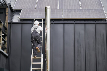 Builder climbing up the ladder while installing photovoltaic solar panels on roof of house. Back...