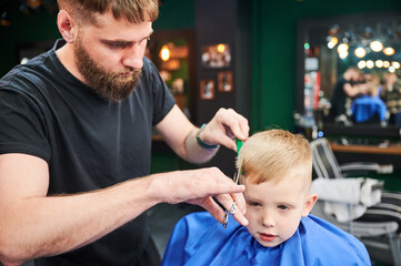 Man barber cutting little boy's hair using comb and scissors. Child getting haircut from adult male, likely barber. Professional hairdresser and cute client at modern barbershop.