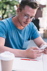 Intelligent male student using cell smartphone phone for research information about delivery service sitting at table in street cafeteria, handsome man in optical eyewear chatting on smartphone