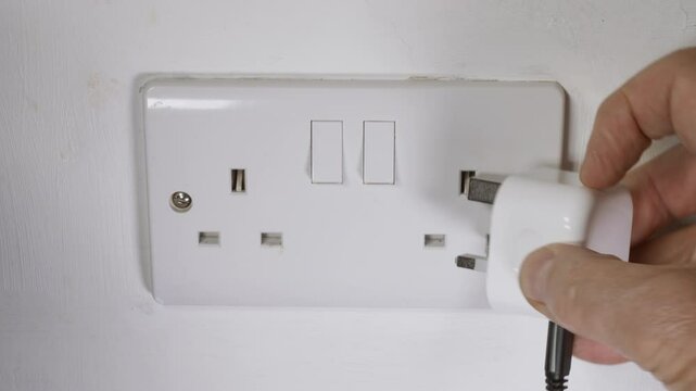 Closeup of man&rsquo;s hand flicking up the three pins of a UK British travel plug with attached cable or cord, then inserting it into a double wall socket and switching the power on.