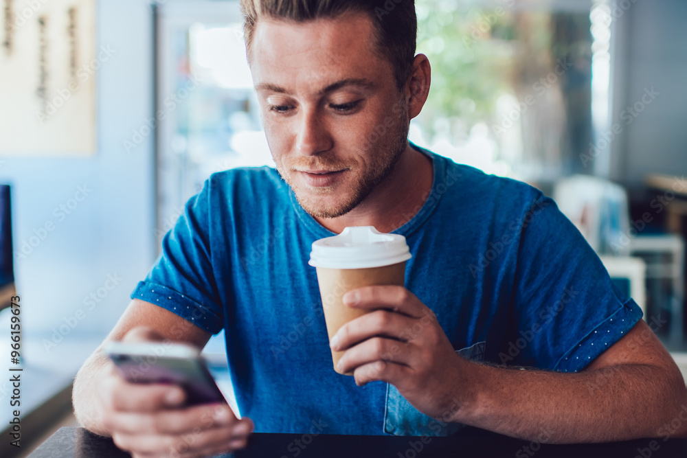 Poster handsome hipster guy holding takeaway cup with caffeine beverage and checking email messages via cel