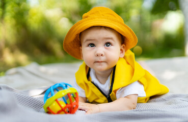 Baby in a yellow hat playing with a colorful ball outdoors. A curious baby wearing a yellow hat and vest explores a colorful ball in a bright outdoor setting surrounded by nature.