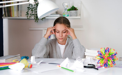 An upset tired child sits at table and does homework among stack of books and scattered toys....