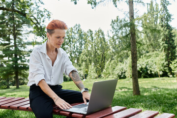A teacher sits on a bench, working on a laptop in a lush green campus setting.