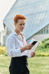 A teacher with red hair checks her phone while holding books on campus.