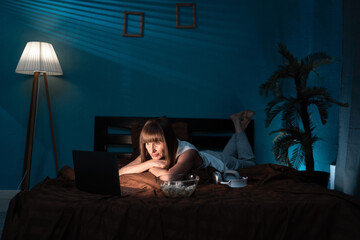 Middle aged woman lying in bed at night watching an interesting movie and eating popcorn