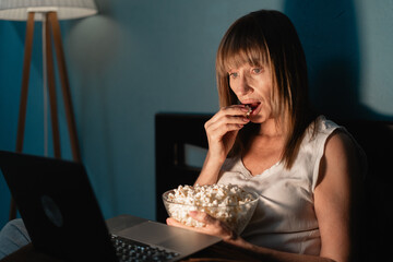 Mature cute woman eating popcorn while sitting on bed under blanket and watching movie