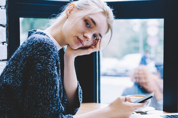 Blonde hair female sitting with smartphone device in hands at the table with open laptop...