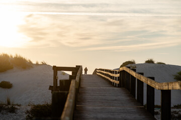 Fototapeta premium A man watches the sunset from the back of a wooden boardwalk, as the golden light of the setting sun creates a serene and peaceful atmosphere, with sand dunes lining the horizon. Costa Nova, Portugal.