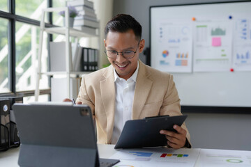 Fototapeta premium Smiling asian businessman analyzing documents and working on digital tablet in modern office