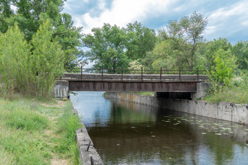 Fototapeta premium Iron concrete bridge over the river canal. Reinforced pedestrian from one bank of water to other. Picturesque trees and bushes grow along the support slabs. Empty footbridge of bridge-canal with pier.