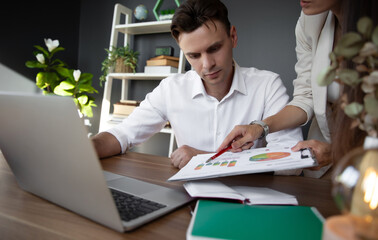 Naklejka premium Man attentively reviews a business chart as colleague points details with a pen. The modern office setup, with laptop and documents, reflects focus, analysis, and teamwork in professional environment