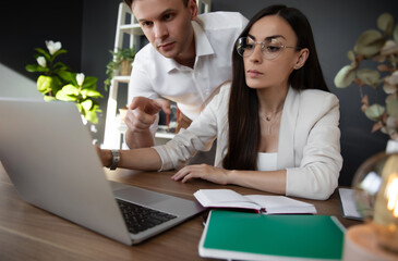 Woman works on laptop in modern office while a colleague gestures, discussing something important. The organized workspace and focused expressions convey collaboration, analysis, and productivity