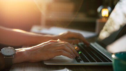 Close-up of hands typing on laptop keyboard, bathed in warm sunlight. The soft glow and gentle...