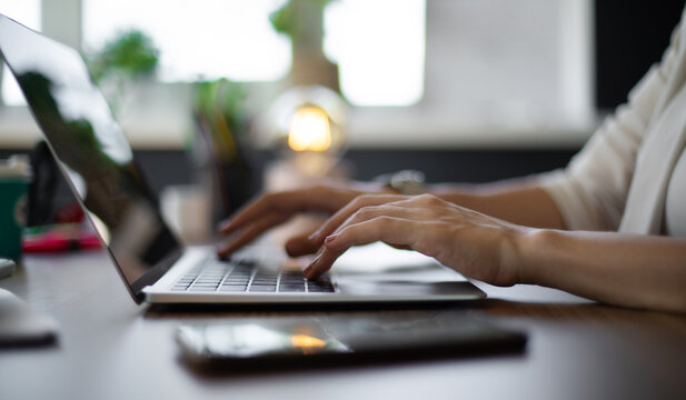 Close-up of female typing on a laptop in a well-lit workspace, with a smartphone resting on the desk. The background features plants and soft lighting, creating a productive and business atmosphere.