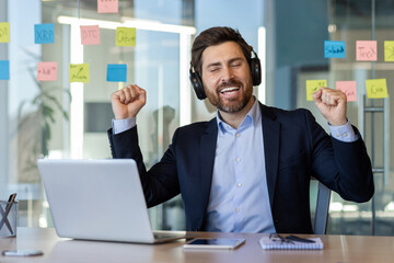 Cheerful businessman expressing happiness listening to music on headphones. Dressed in formal suit, working on laptop with sticky notes on glass wall. Capturing productivity, success, and positive