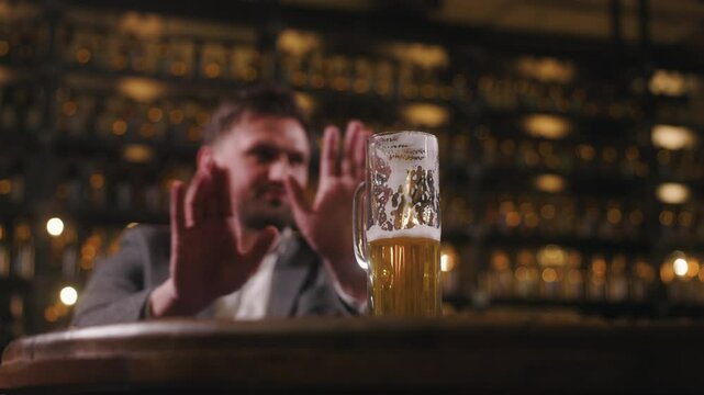 A man sitting in a bar or pub is refusing a glass of beer by raising his hand in a gesture of denial. The focus is on the half-full beer glass, with foam marks visible on the inside.