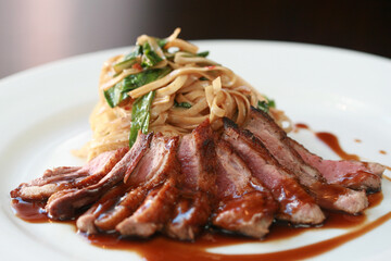 Sliced duck breast served with bok choy and noodles drizzled with sauce on white plate