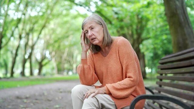 A sick senior female with gray hair suffering from a headache while sitting on a bench on the street in an urban city park. Upset overtired elderly woman massages and rubs her forehead and temples
