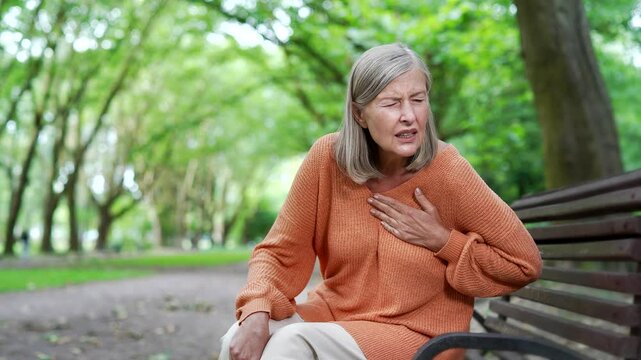 Sick senior female with gray hair suffering from chest tension, heart attack sitting on bench in city park. Elderly woman feels anxious, holding her chest with her hand, needs urgent medical attention